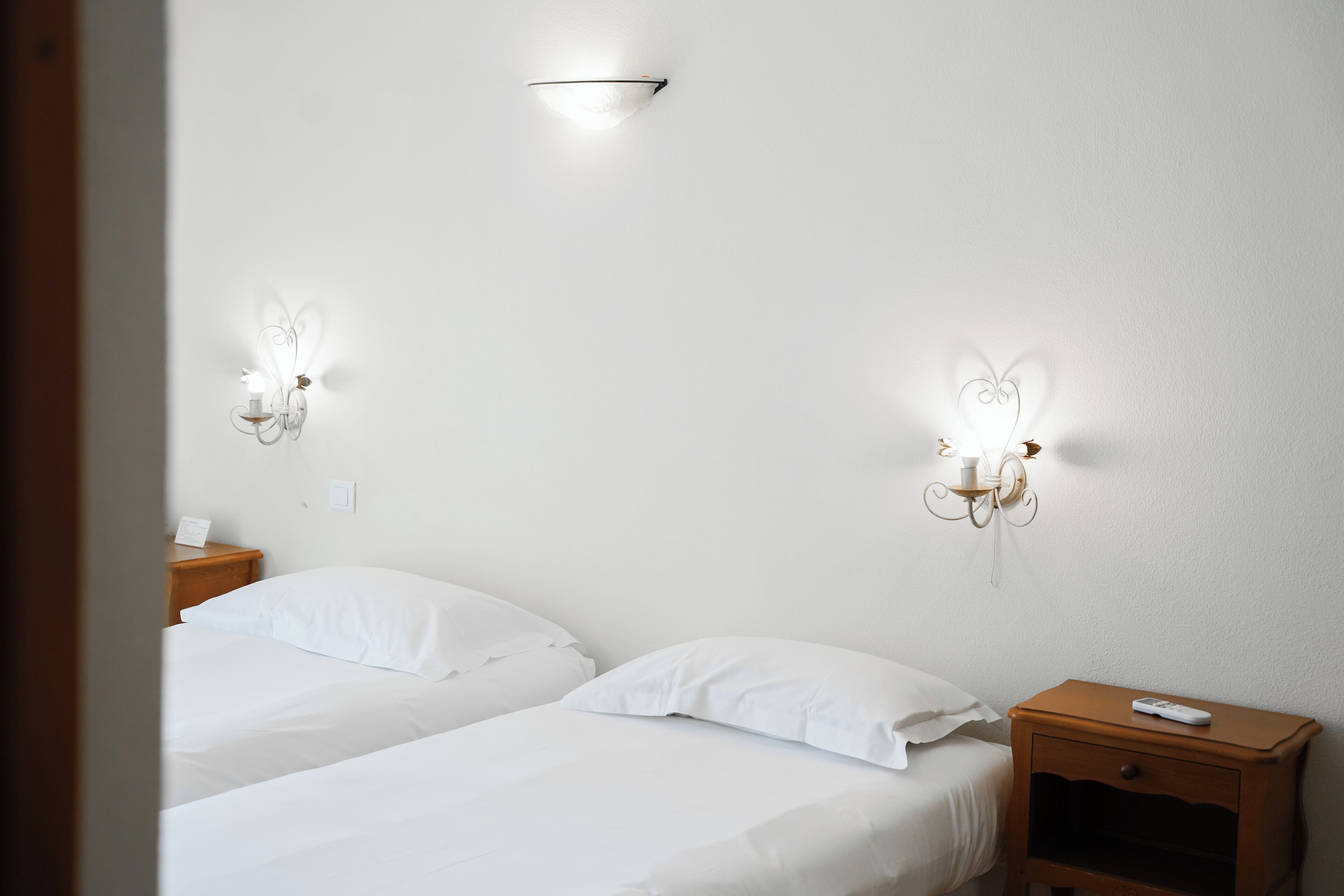 A hotel room with two single beds, a bedside table with a remote control, and wall-mounted light fixtures.