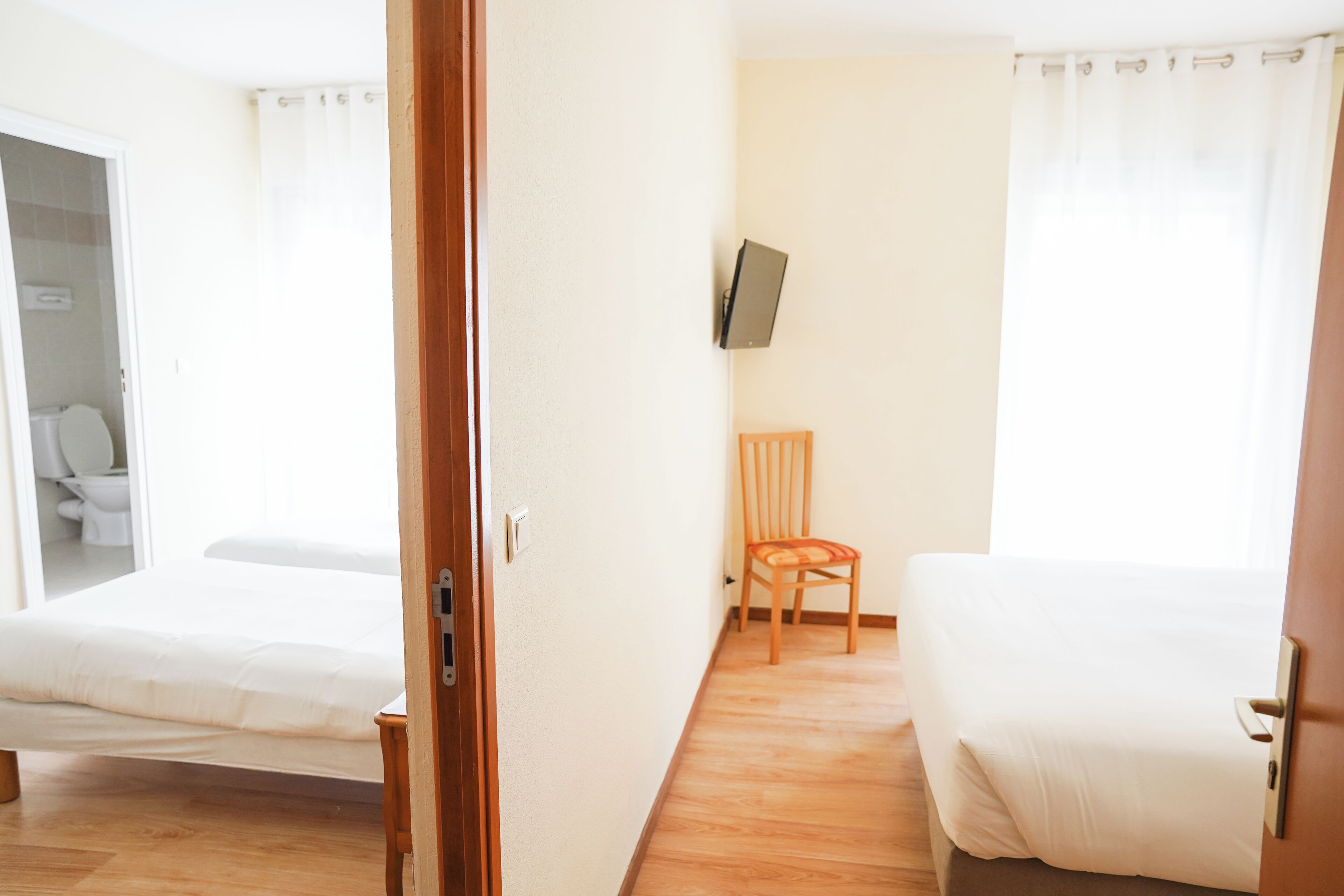 Ein Hotelzimmer mit zwei Betten, einem Holzstuhl, einem an der Wand befestigten Fernseher und Holzboden.