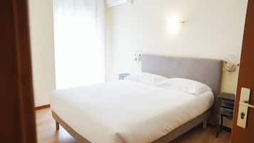 Una cama bien tendida en una habitación con piso de madera, una mesita de noche, un aire acondicionado de pared y una lámpara de pared.