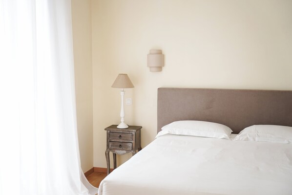 A neatly made bed with white linens, a bedside lamp, and a small wooden nightstand with drawers. - Hôtel Castellu (Lucciana)