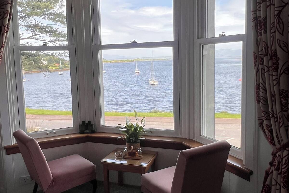 A room with a bay window overlooking a body of water, two pink chairs, a small table with a plant, and a view of sailboats.