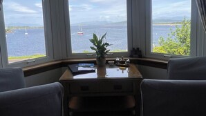 A room with a bay window overlooking a body of water, a small table with a plant, and two chairs.