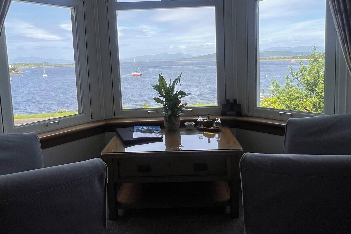 A room with a bay window overlooking a body of water, a small table with a plant, and two chairs.
