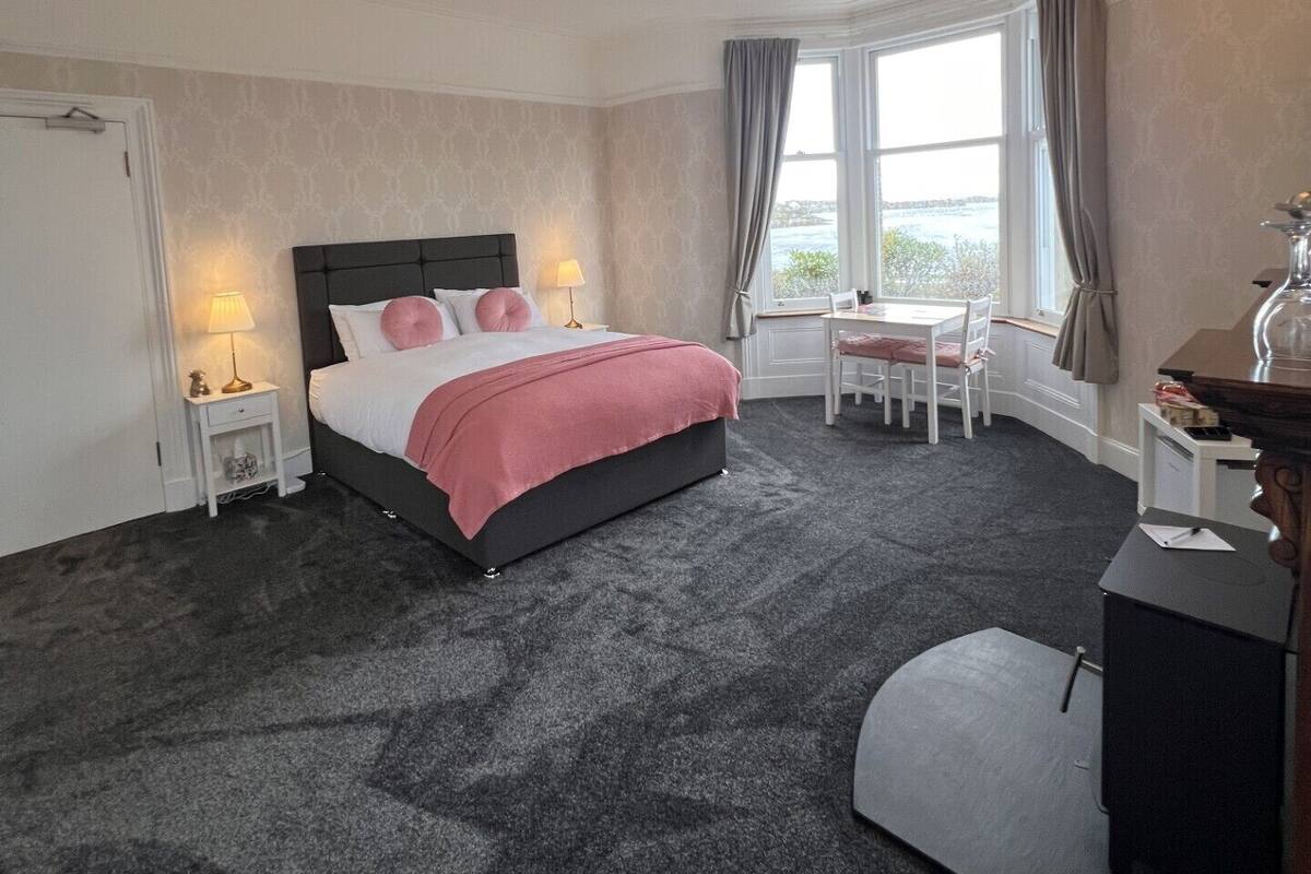 A hotel room with a bed, bedside tables, a desk, a chair, and a view of the sea.