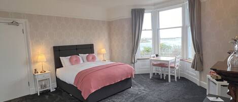 A hotel room with a bed, bedside tables, a desk, a chair, and a view of the sea.