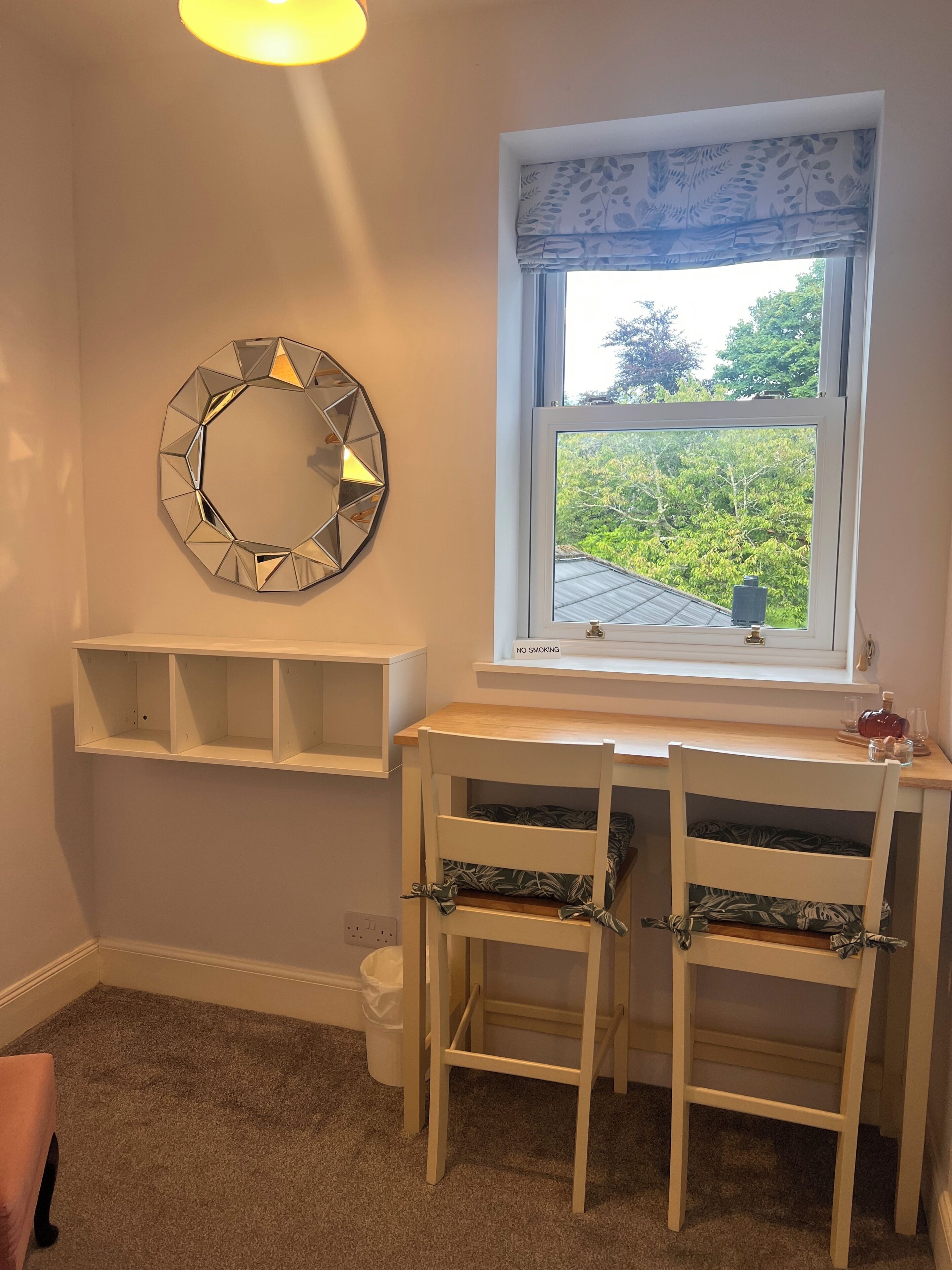 A room with a desk, two chairs, a window with a view of greenery, a wall-mounted mirror, and a 'NO SMOKING' sign.