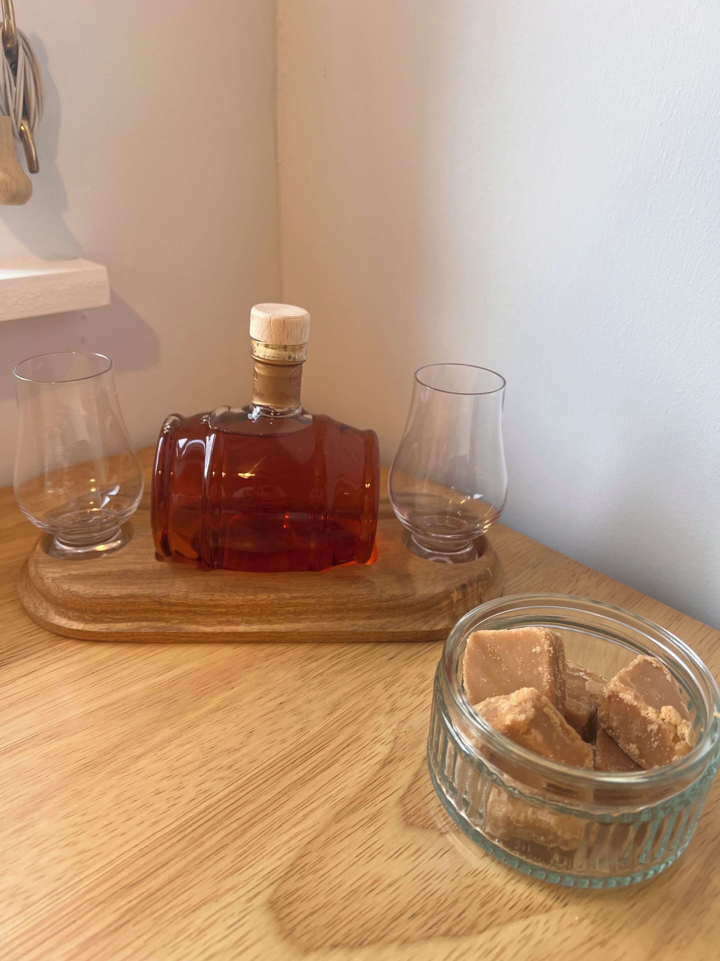 A wooden table with a bottle of amber-colored liquor, two empty glasses, and a small glass jar with a brown substance.