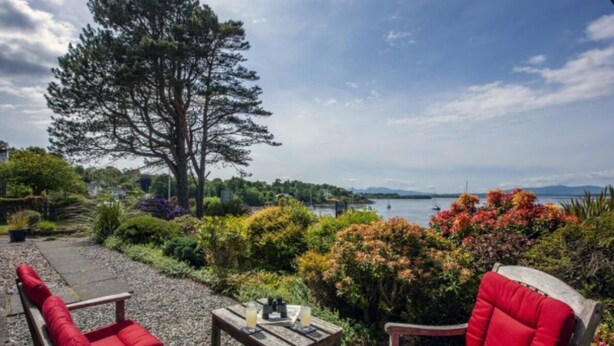 A patio with two red cushioned chairs and a small table, overlooking a lake with mountains in the distance.
