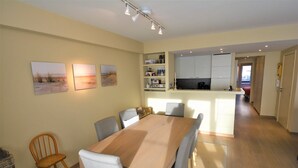 Un coin repas avec une table et des chaises en bois, une cuisine avec des meubles blancs et un couloir menant à une autre pièce.