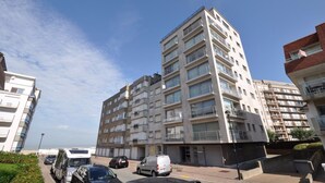 Um edifício residencial com vários andares, varandas, um caminho pavimentado e veículos estacionados.