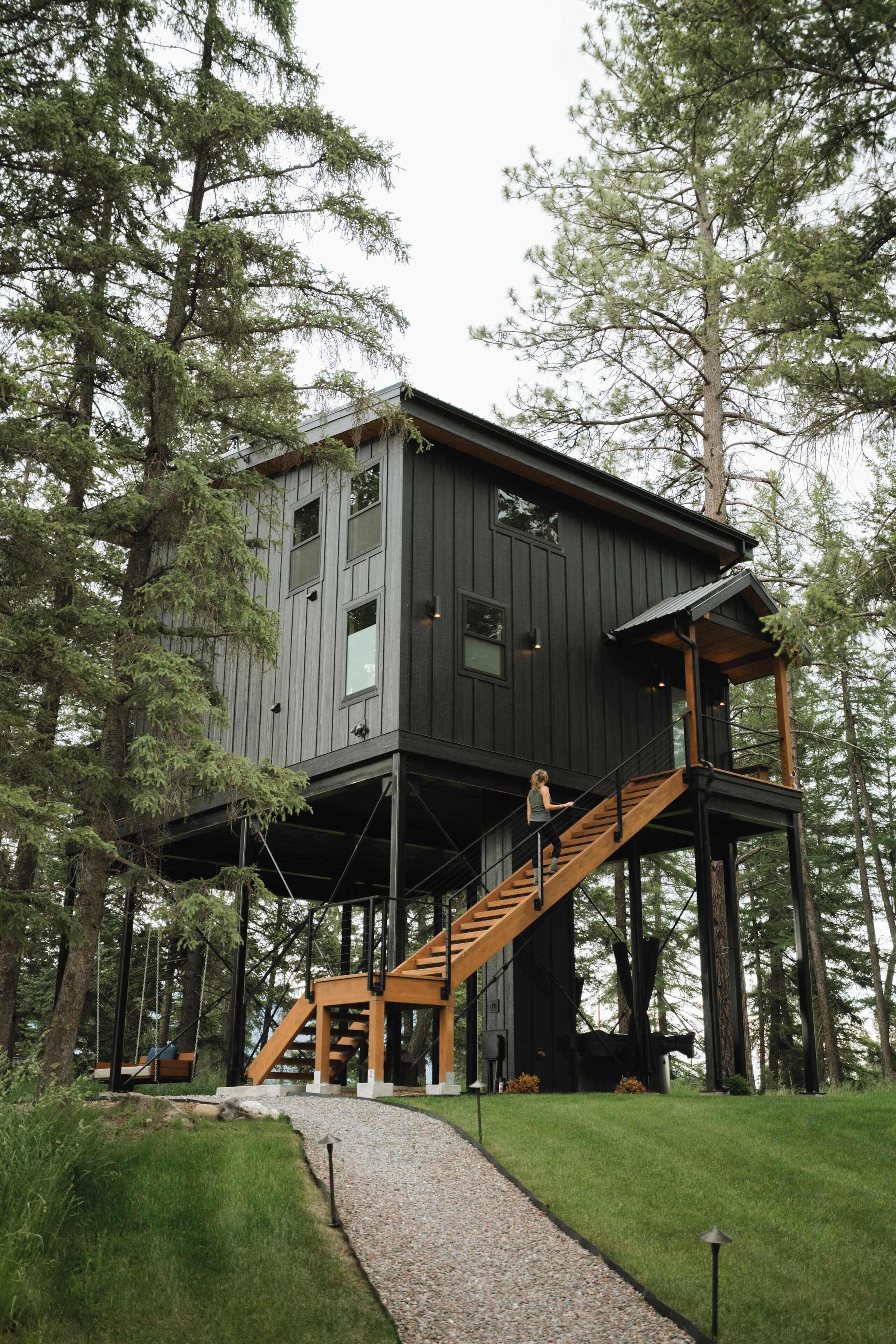 A modern wooden cabin with a staircase leading to a second level, surrounded by trees.