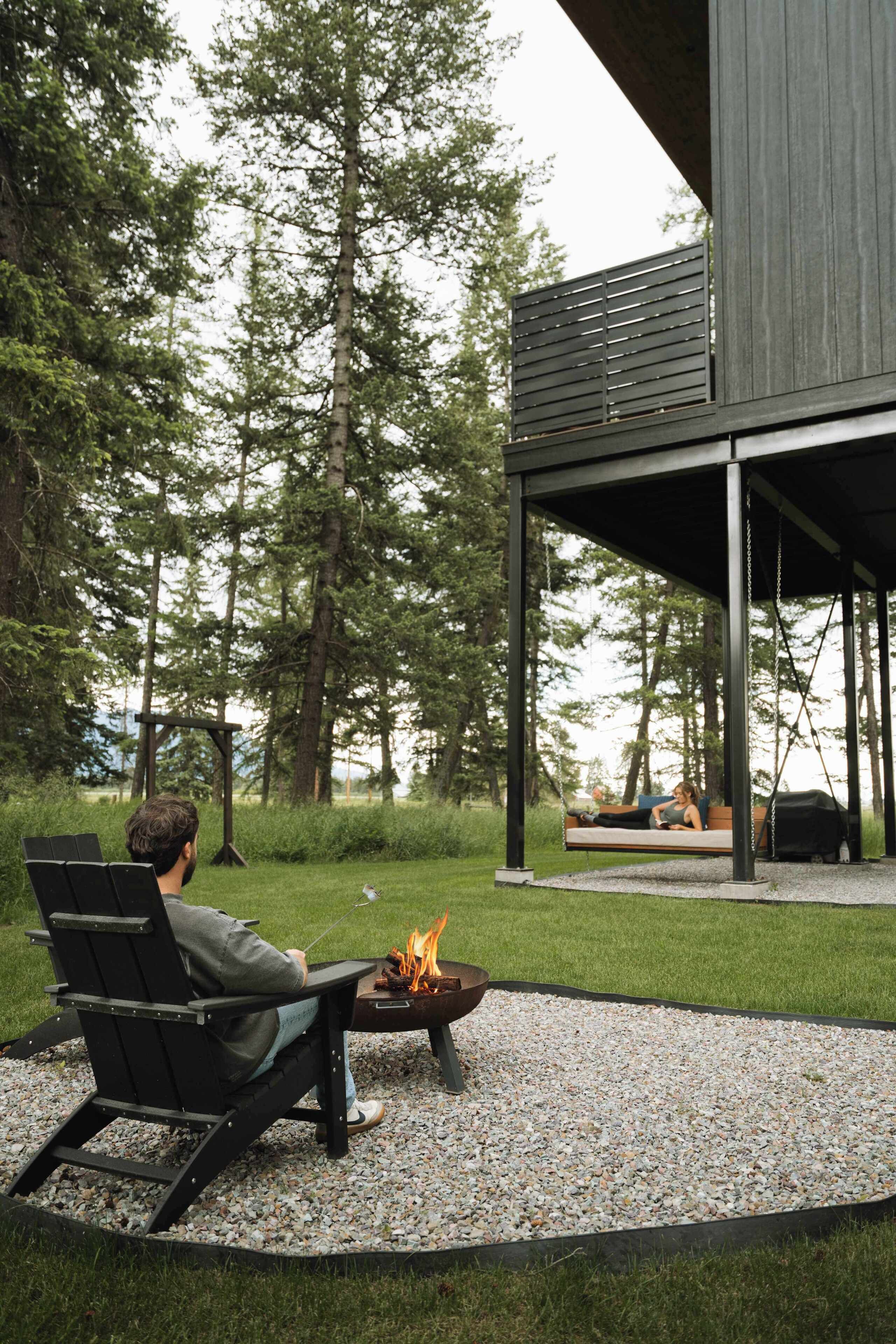 A person relaxing on a chair by a fire pit with a view of a forest and a modern building.