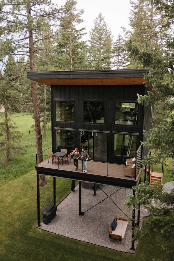 Una cabaña moderna con terraza, ventanas amplias y una escalera que lleva a un nivel superior.