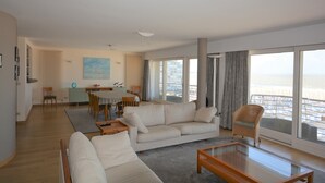 En rummelig opholdsstue med sofa, sofabord og spiseplads. Store vinduer giver udsigt til stranden.
