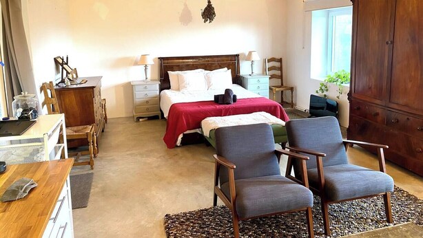 Une chambre avec un plafond en bois, un lit avec une literie blanche, deux fauteuils, une commode et une fenêtre donnant sur un beau paysage.