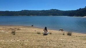 Une femme est assise sur une plage de galets, avec un lac et des collines boisées en arrière-plan.