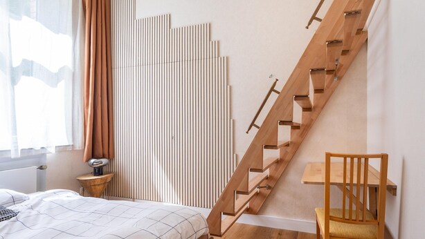 Een slaapkamer met een houten trap, een bed met een zwart-wit geruite sprei, een houten stoel en een nachtkastje met een lamp.