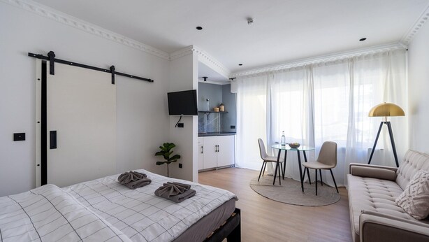 A modern hotel room with a bed, a sofa, a dining area with a table and chairs, a TV, and a kitchen area.