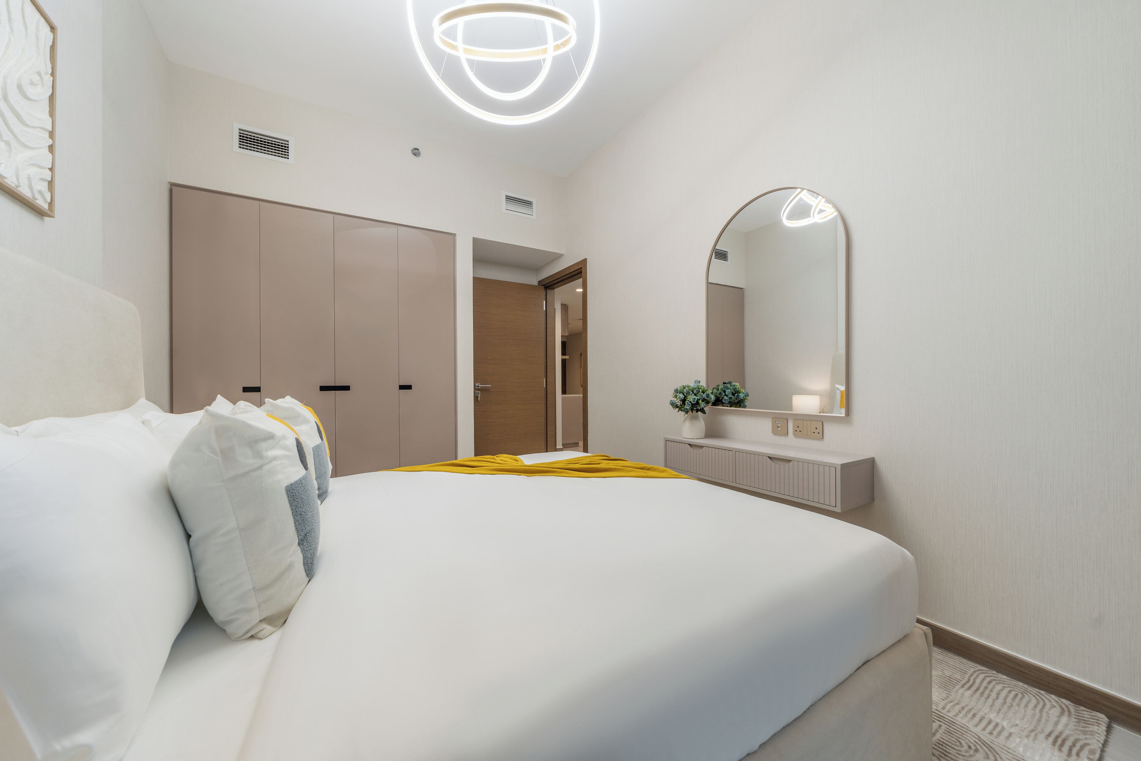 A modern hotel room with a large bed, a bedside mirror, a small table with a vase, and a wardrobe.