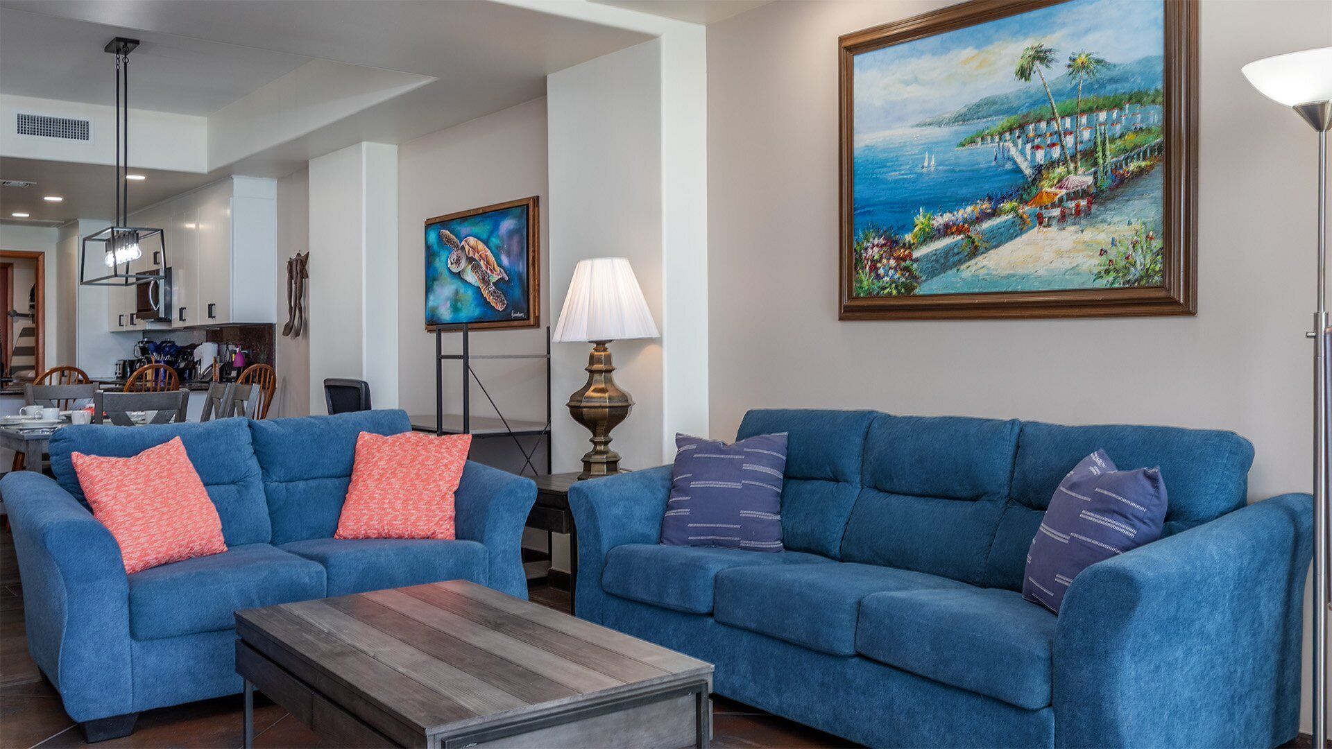 A living room with two blue sofas, a coffee table, a painting of a beach scene, and a lamp.