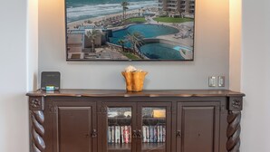 Une chambre d’hôtel dotée d’une grande fenêtre donnant sur une plage et une piscine, d’un meuble en bois contenant des livres et d’un petit haut-parleur posé sur la cheminée.