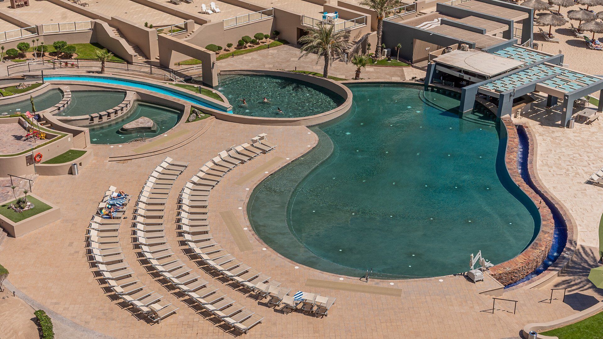 Una amplia piscina con diseño curvo, rodeada de sillas de descanso y una zona cubierta con sombrillas.