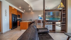 A modern kitchen with a built-in refrigerator, microwave, and coffee maker. There is a dining area with a table and chairs, and a large TV mounted on a wooden shelf.