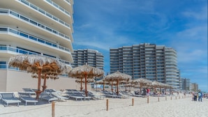 Ein Strand mit Liegestühlen und Reetdach-Sonnenschirmen, umgeben von Hochhäusern.