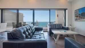 A modern lounge area with leather sofas and a coffee table, large windows with a view of the sea, and a painting on the wall.