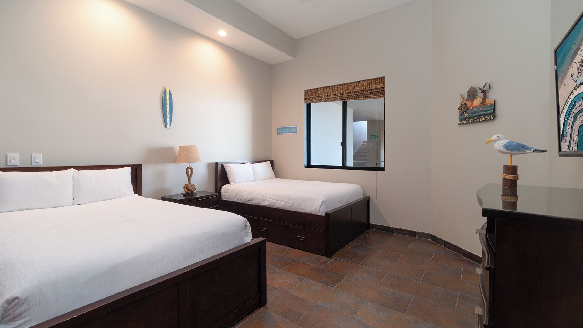 Une chambre d’hôtel avec un grand lit, un plus petit lit, une table de chevet avec une lampe, une commode, un miroir et un mur orné d’une pancarte de bienvenue et d’une décoration représentant un goéland.