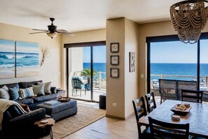 A living room with a sofa, a dining table with chairs, a ceiling fan, and a view of the ocean through large glass doors.