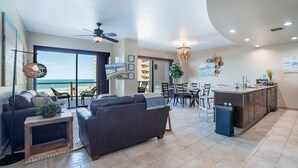 Un vaste espace de vie donnant sur la plage, équipé d’un ventilateur de plafond et comprenant un coin repas avec des chaises et une table.