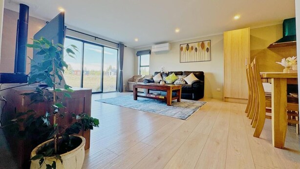 A spacious living room with a large window, a sofa, a coffee table, and a dining area with wooden chairs. - April Three Bedroom Apartment (Lake Tekapo)