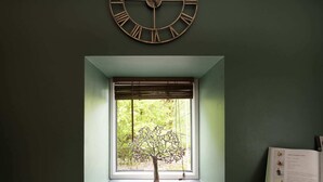A room with a window, a wall clock, a small tree sculpture, and a book on a table.