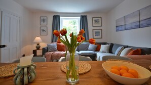 A living room with a wooden table, a vase with orange flowers, a bowl of oranges, and a couch with cushions.
