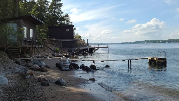 A wooden cabin with a deck, a dock extending into calm water, and a rocky shoreline. - Ref. 534251 (Saltsjö-Boo)