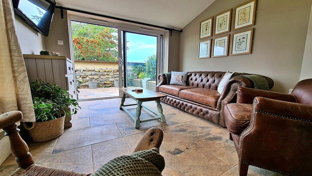 A cozy living room with a brown leather sofa, a wooden chair, a small table, and a large window with a view of a garden. - Cottage in Glaisdale by North Yorkshire Moors (Whitby)