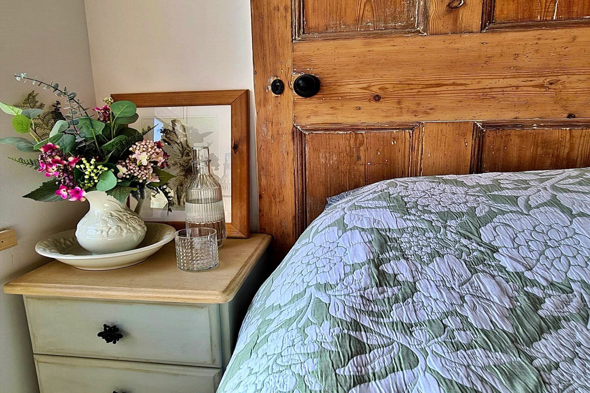 Une chambre à coucher avec une couvre-lit à motifs floraux, une porte en bois, une table de chevet ornée d’un vase de fleurs et un tableau encadré.