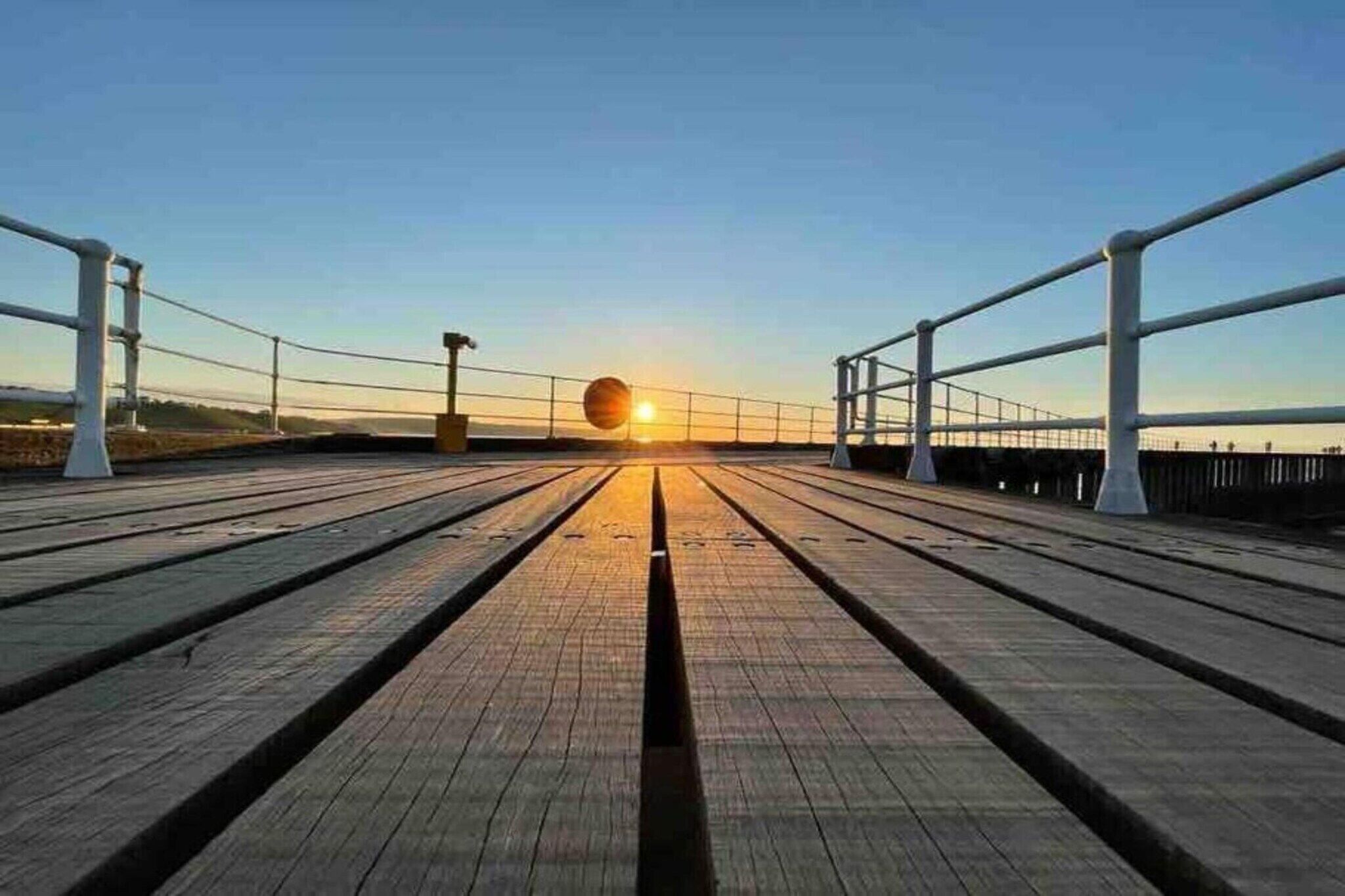 Um deck de madeira com guarda-corpos de metal e vista para o pôr do sol.