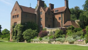 Un grand bâtiment historique en briques, doté de nombreuses cheminées, entouré de jardins bien entretenus et surplombé d’un ciel dégagé.