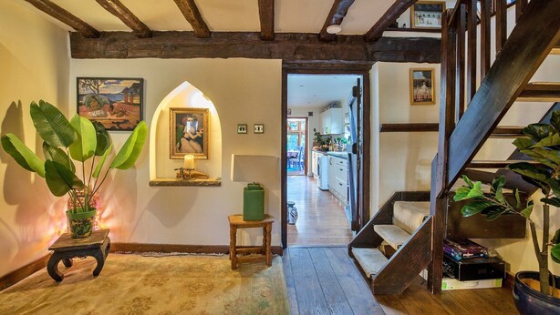 Un couloir avec des poutres en bois, une plante verte, un tableau encadré et un escalier menant à une autre pièce.