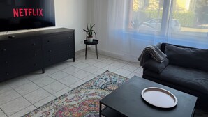 Ein Wohnzimmer mit Fernseher, der 'NETFLIX' anzeigt, einer dunklen Couch, einem kleinen Tisch mit Tablett, einem gemusterten Teppich und einem Blumentopf.