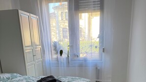 Ein Schlafzimmer mit Bett, Schrank, einem Fenster mit durchsichtigen Vorhängen und Blick ins Freie.