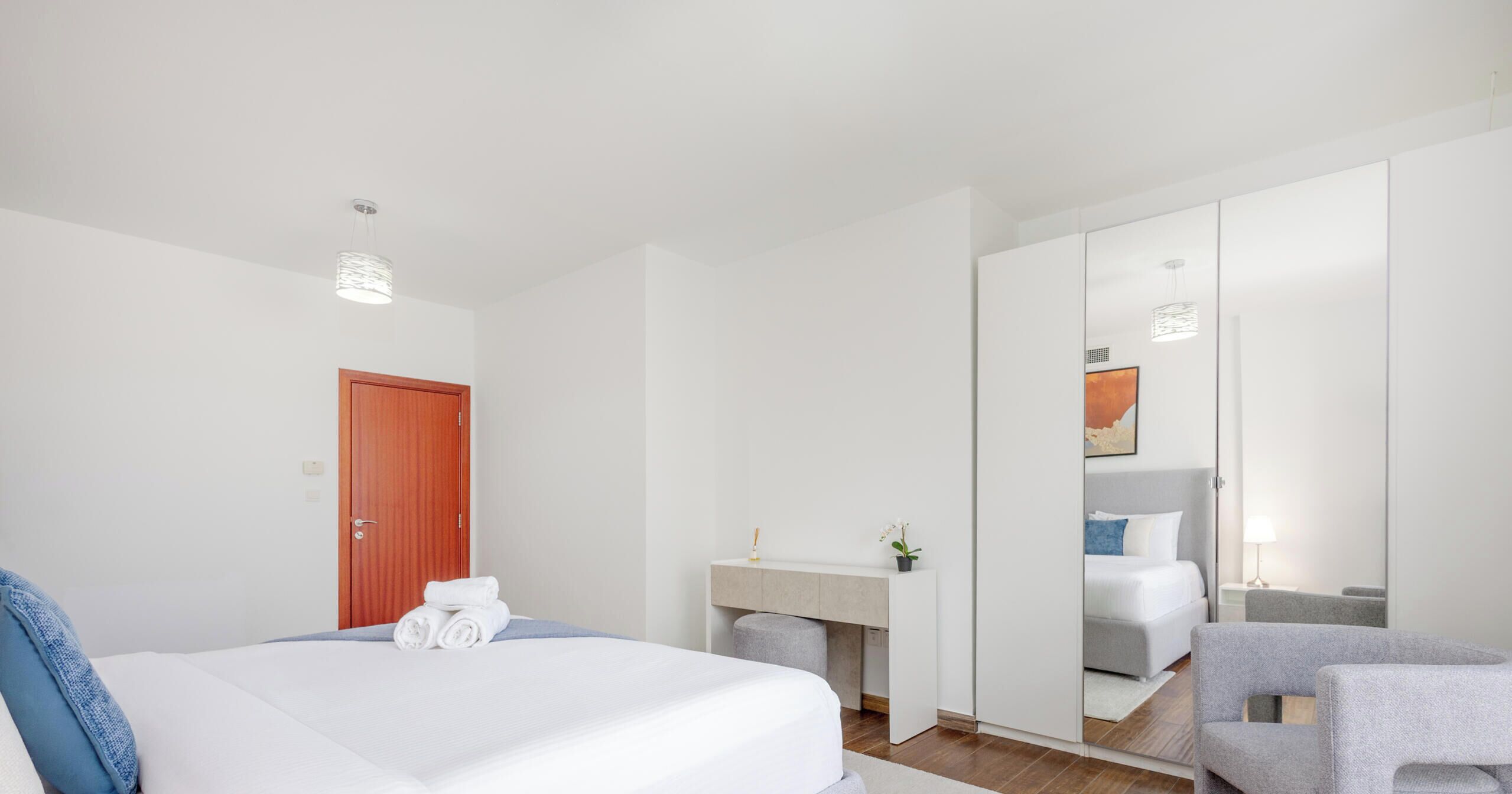 A modern hotel room with a large bed, a wooden door, a bedside table with a lamp, a mirror, and a seating area with a chair.
