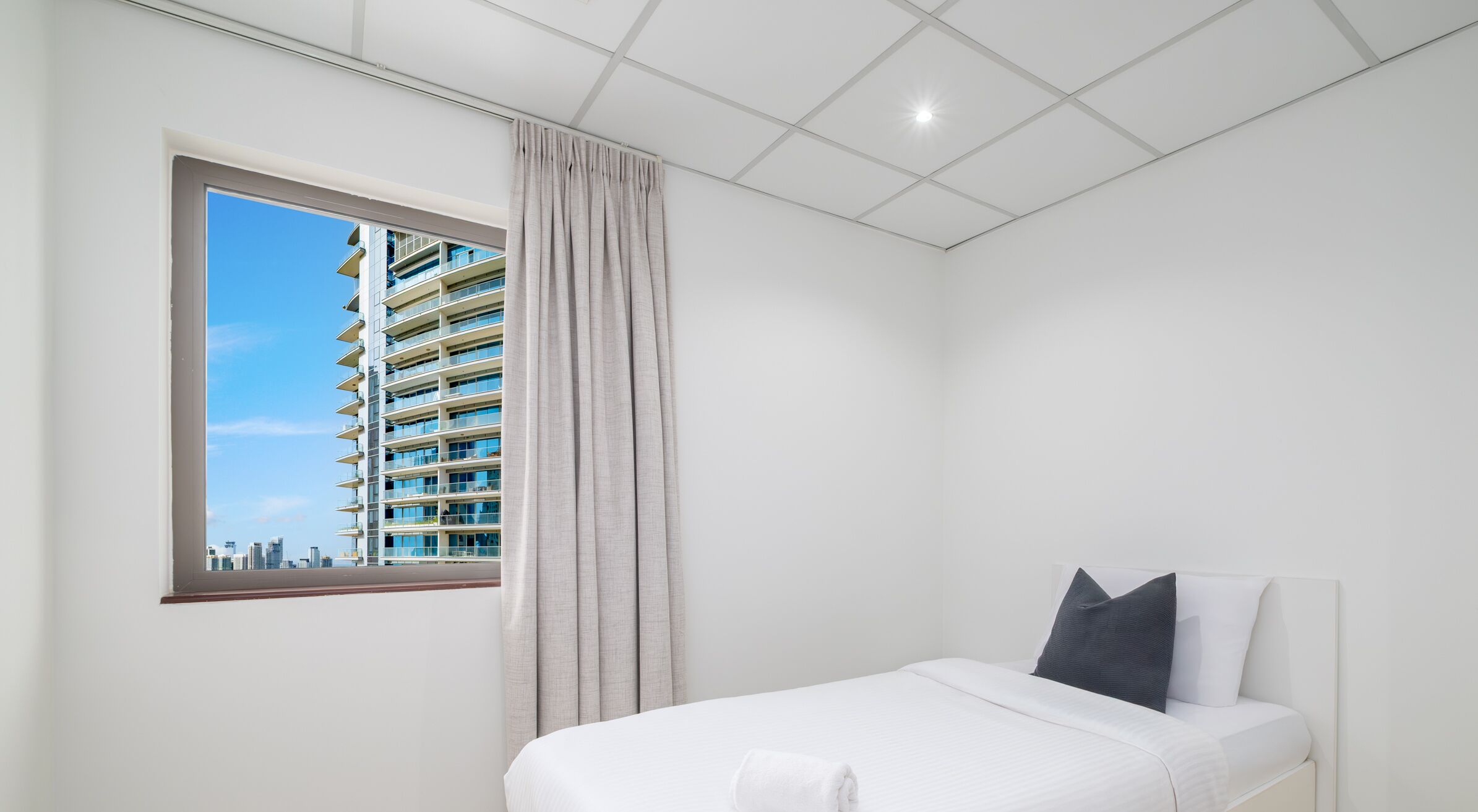 A modern hotel room with a large window, a bed with white linens, and a view of a high-rise building.