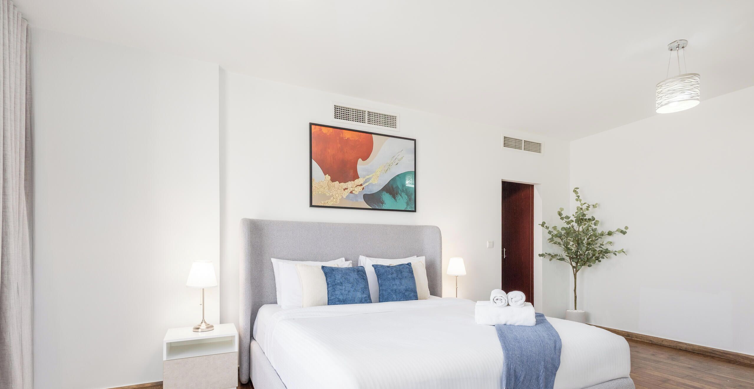 A modern hotel room with a large bed, a headboard, a wall-mounted abstract painting, a bedside table with a lamp, and a potted plant.
