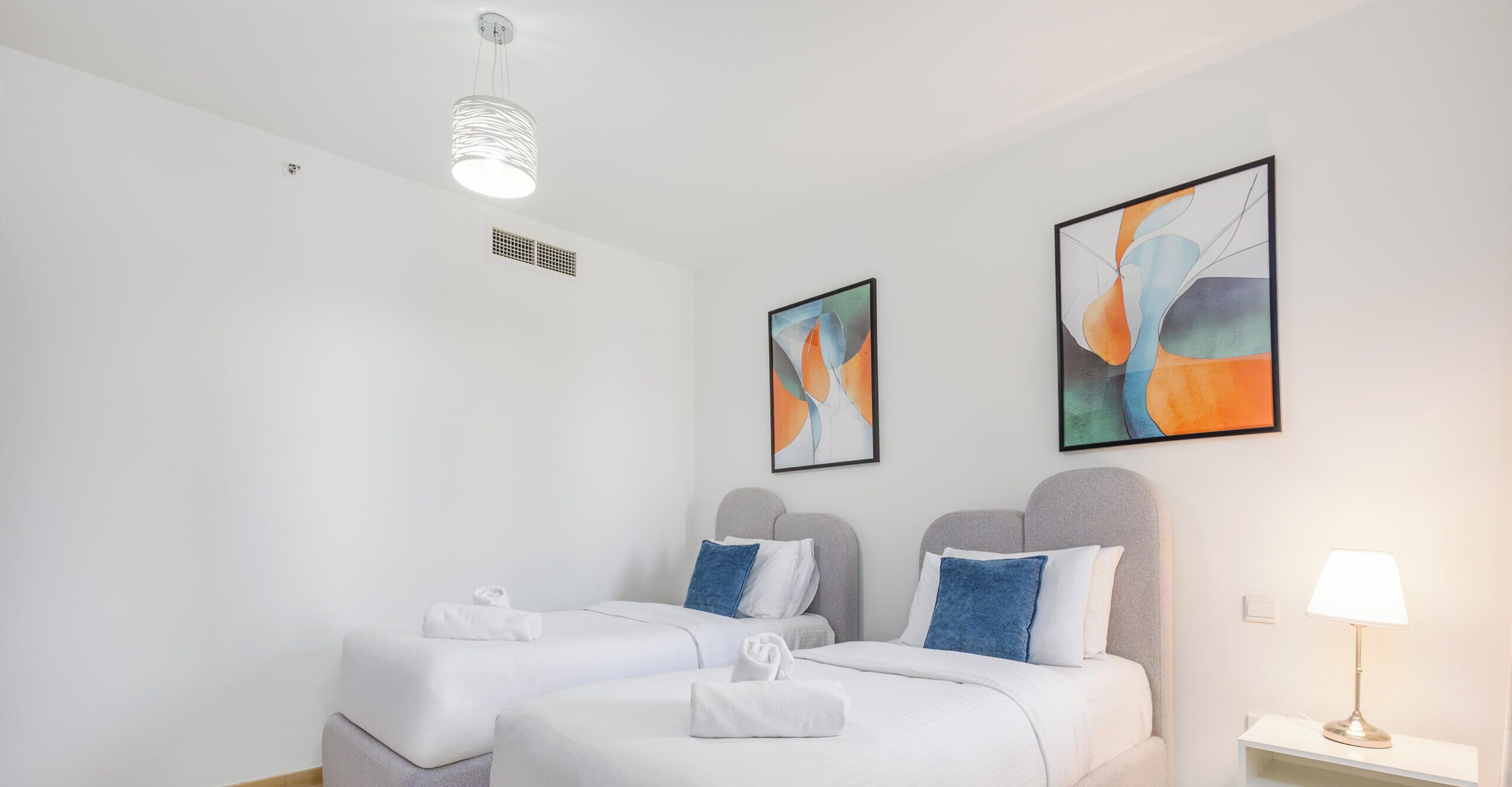 A hotel room with two beds, a ceiling light, and two abstract paintings on the wall.
