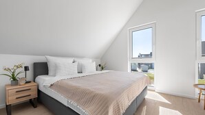 A modern bedroom with a sloped ceiling, a large window, a bed with white bedding, a bedside table with a lamp, and a small potted plant. - Haus Fabelhaft - Terrace, 2 Bathrooms, Fireplace, Infrared Sauna, SodaStream (Olpenitz)
