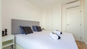 Una cama bien tendida con cabecera gris, paredes blancas y un armario blanco con puerta.