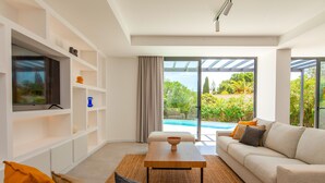 A modern living room with a flat-screen TV, white shelving, a beige sofa, and a view of a swimming pool. - Villa Farol Boutique (Lagoa)